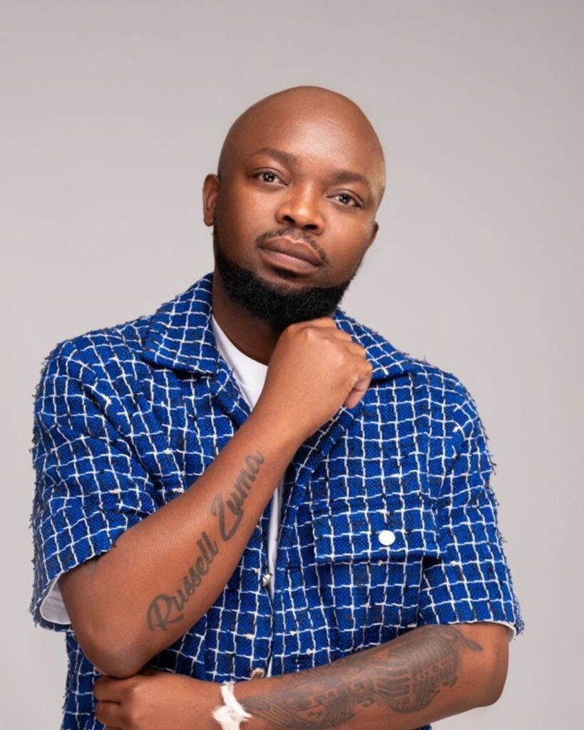 Sunset Jazz Melodies Artist from the Lineup Russell Zuma a bald man dressed in a blue and white checkered shirt. He will be performing in 6 Decemeber at the SUnset Jazz Melodies festival at Albert Falls Dam